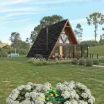 Projeto de chalé de madeira pré-fabricado. O chalé tem uma arquitetura moderna, com uma varanda cercada com aço escuro. Ao redor dele um gramado com hortências, sugerindo uma casa de campo.